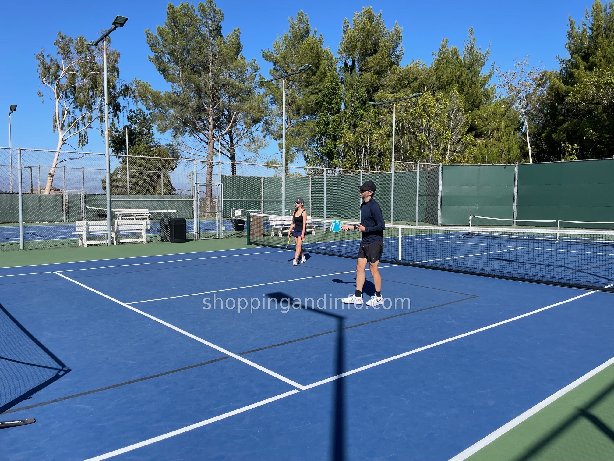 Two people rallying on a blue hard tennis court backed by trees